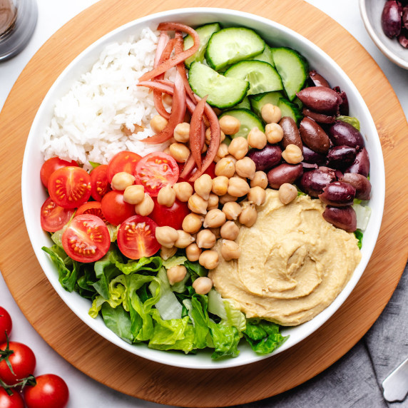 A bowl with white rice, cherry tomatoes, cucumbers, lettuce, hummus, olives, chickpeas, and onions.
