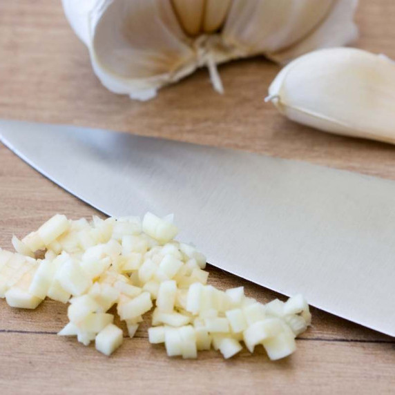 Minced garlic sits beside a knife on a wooden cutting board, with cloves of garlic in the background