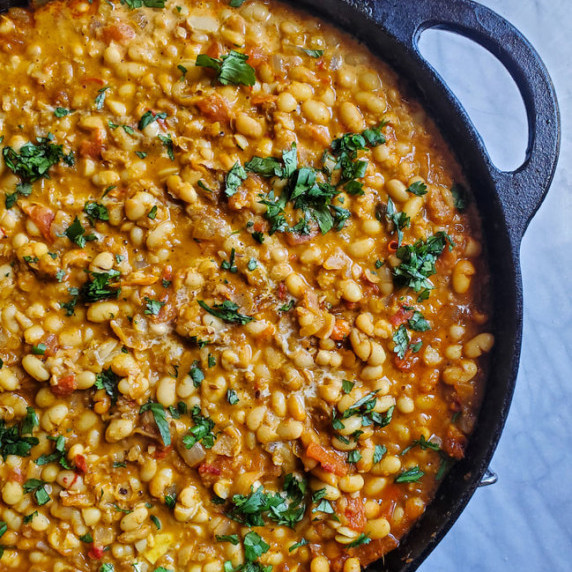 A skillet of navy beans in a robustly flavoured curry sauce. Set on a marble background.