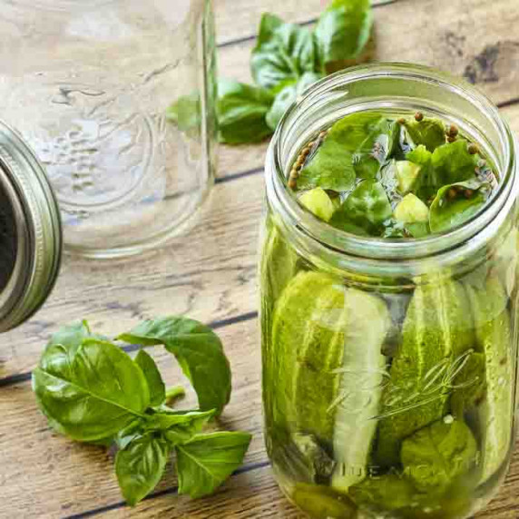 Side shot of pickles in a mason jar on a wooden table with basil and an empty mason jar on the side.