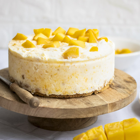 Side shot of the mango cheesecake on a raised wooden cake platter with a knife and mango on the side