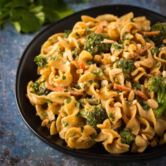 Close up angled shot of noodles and broccoli garnished with chopped peanuts on a black plate.