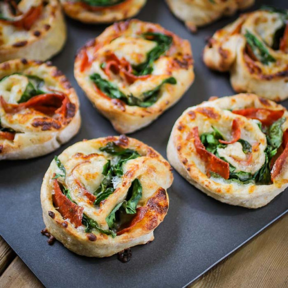 Angled close up shot of pepperoni pinwheels arranged on a metal baking sheet.