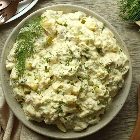 Potato salad with dill pickles in a tan speckled bowl on wooden surface.