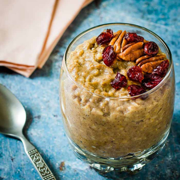 Side shot of overnight oats in a clear glass cup garnished with pecans and cranberries on a blue spe