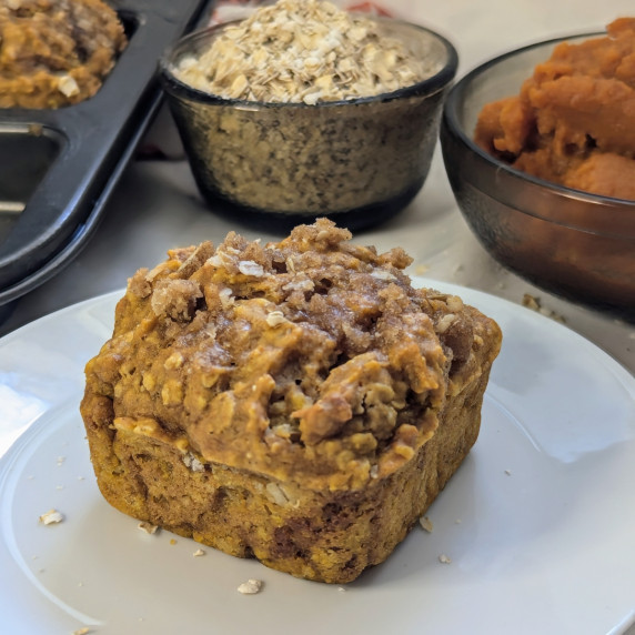 A pumpkin spice oatmeal muffin with a bowl of oats and a bowl of pumpkin puree in the background.