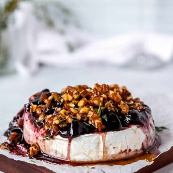 A wheel of baked brie with jam and nuts on top, garnished with thyme, on a wooden serving board.