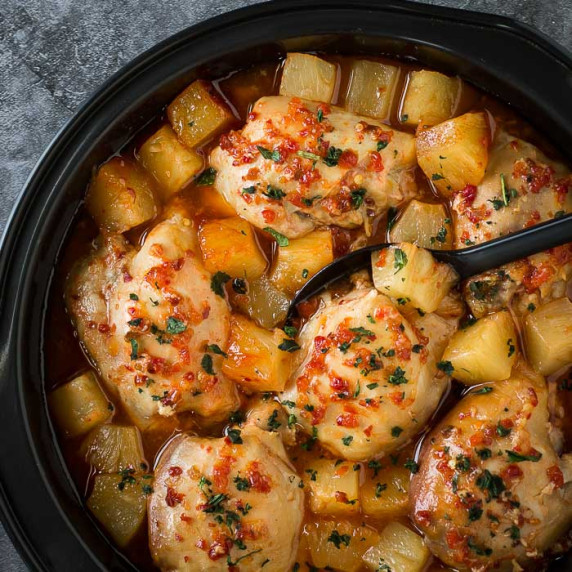 Overhead shot of Hawaiian chicken in the crockpot with a black spoon on a grey marble counter.