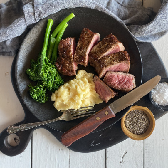 Sliced steak, potatoes and brocollini on a plate with a steak knife, fork and salt and pepper 