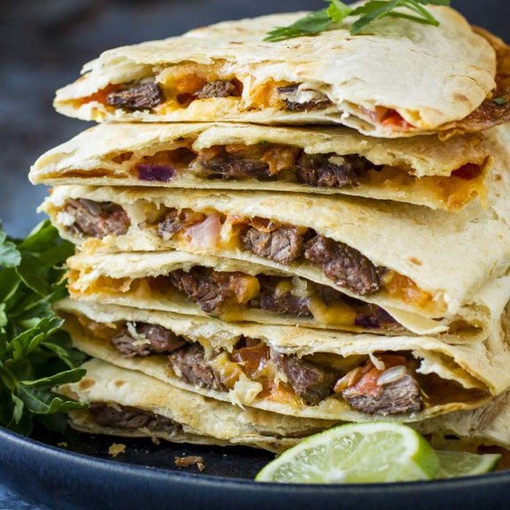Side shot of multiple steak quesadillas stacked on a black plate with fresh herbs and lime wedges.