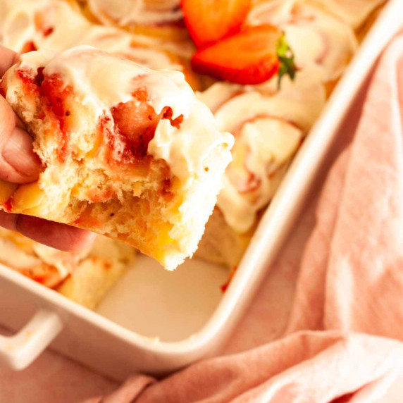 A hand holding a strawberry cinnamon roll over a baking pan.