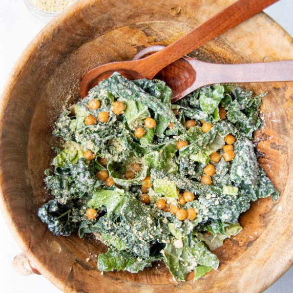 Large wooden bowl filled with kale caesar salad topped with crunchy chickpeas and hempseed parmesan.