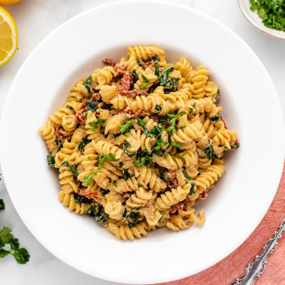 Tahini pasta with sun-dried tomatoes, kale, and chopped parsely in a white bowl.