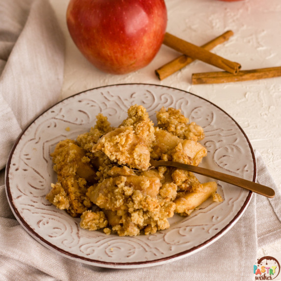 vegan apple crisp on a plate with apples and cinnamon sticks in the background